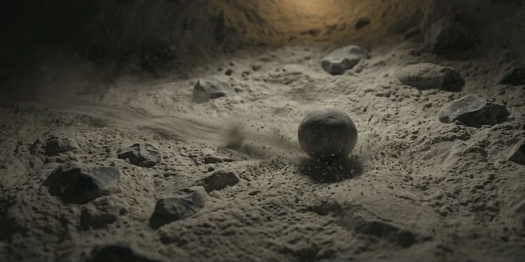 A pebble rolling across the earth floor of a tunnel, trailing dust, in faint amber light.